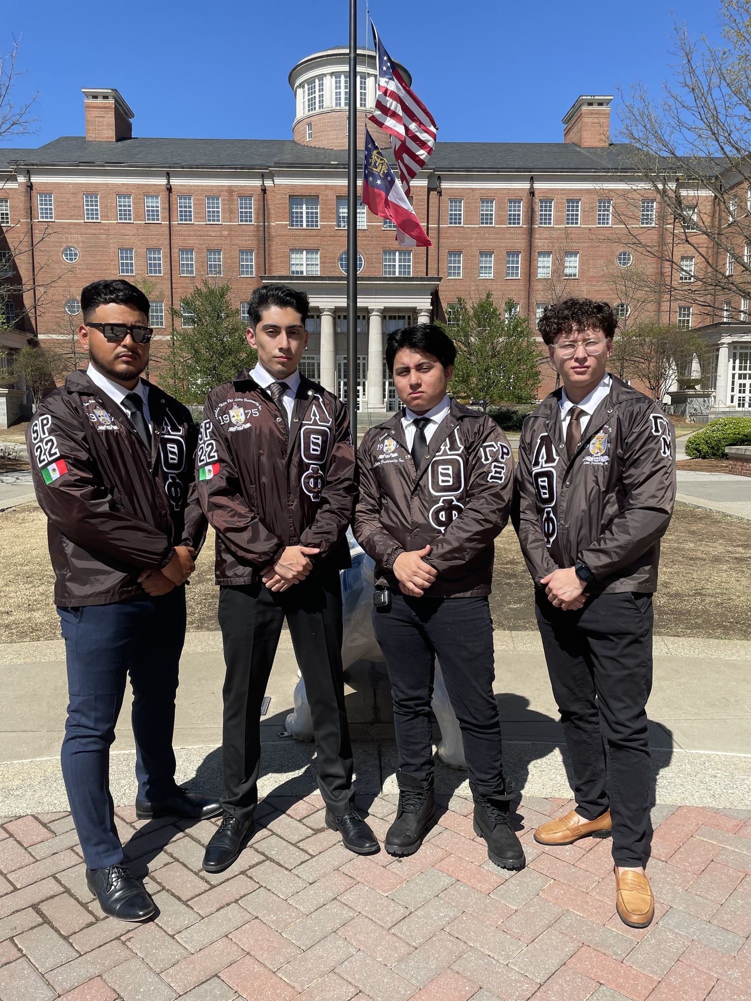 Four brothers in fraternity jackets standing in a line outside a brick campus building, flags flying behind them.
