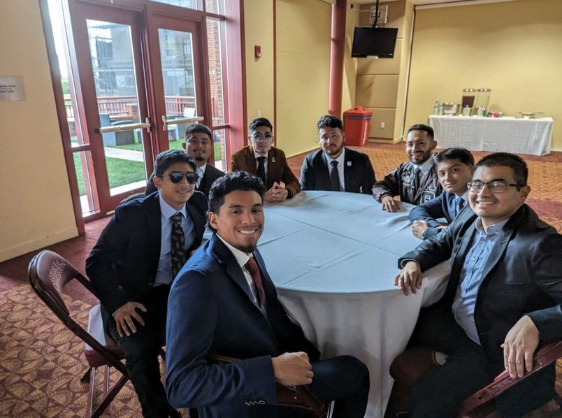 Eight brothers in suits seated around a round table at a chapter event.